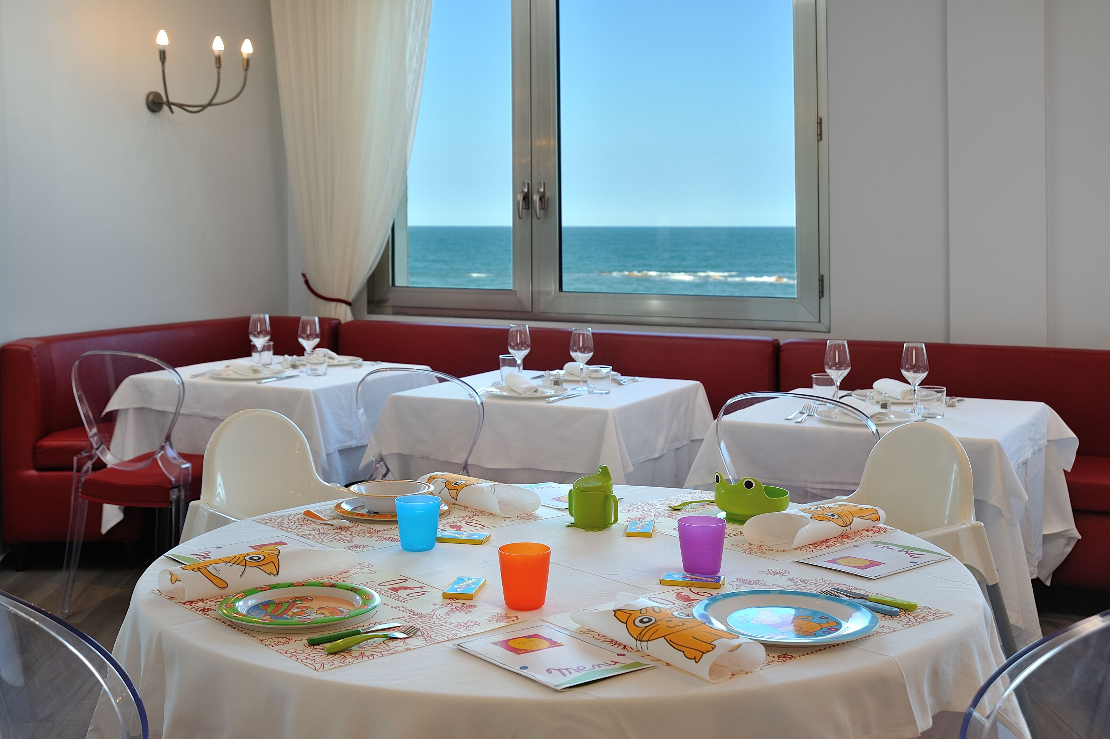 A dining area with tables set for a meal, a view of the ocean through large windows, and a red bench along the wall.