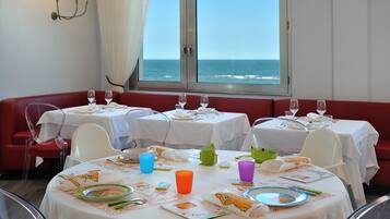 A dining area with tables set for a meal, a view of the ocean through large windows, and a red bench along the wall.
