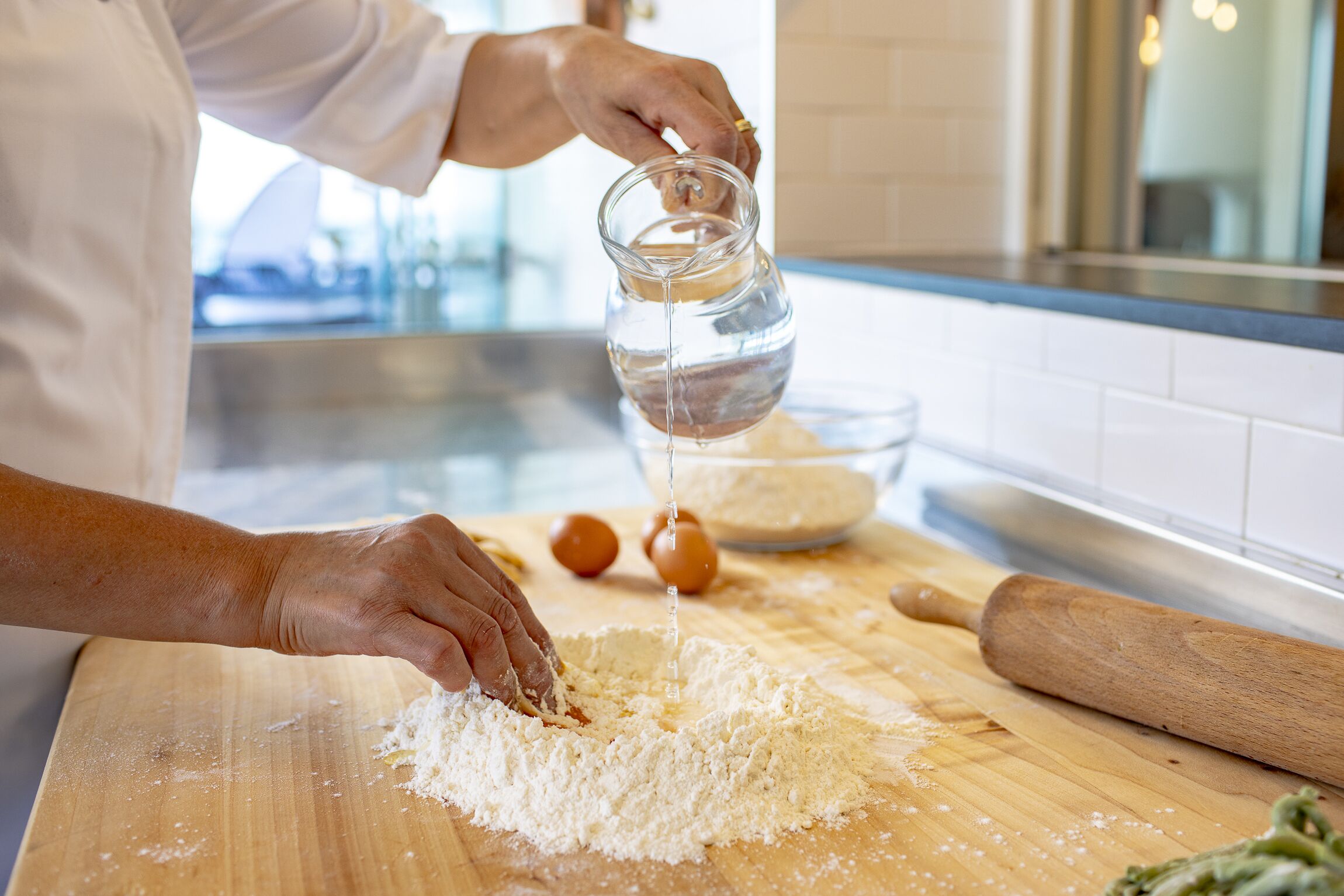 A person pouring water into a bowl of flour on a wooden cutting board in a kitchen.