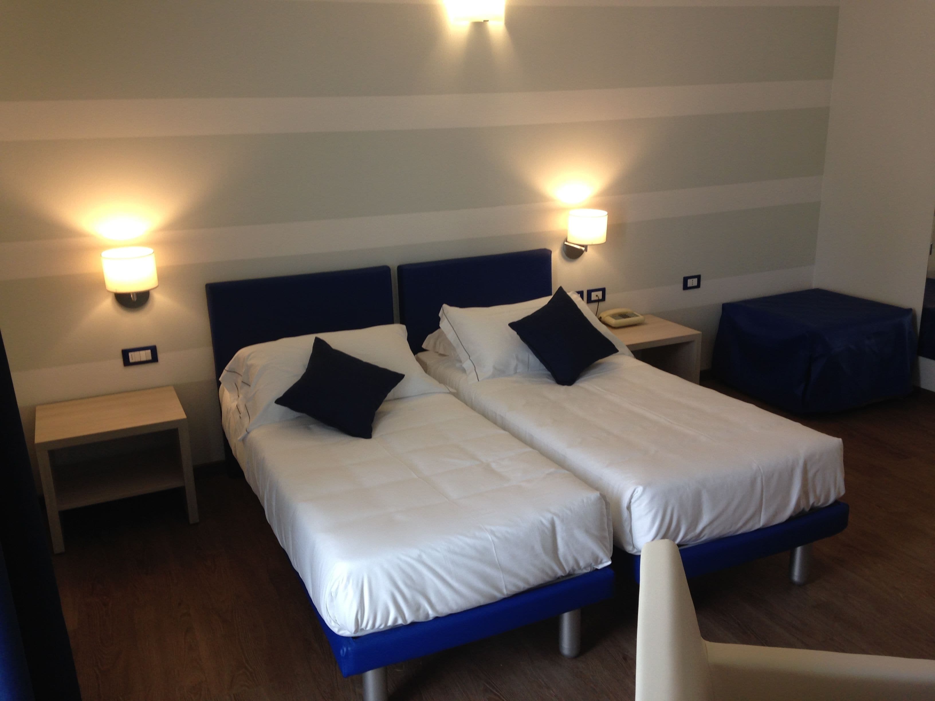 Habitación de hotel con dos camas, cada una con cabecera azul y ropa de cama blanca. Hay lámparas de noche, mesitas de noche y un pequeño otoman azul.