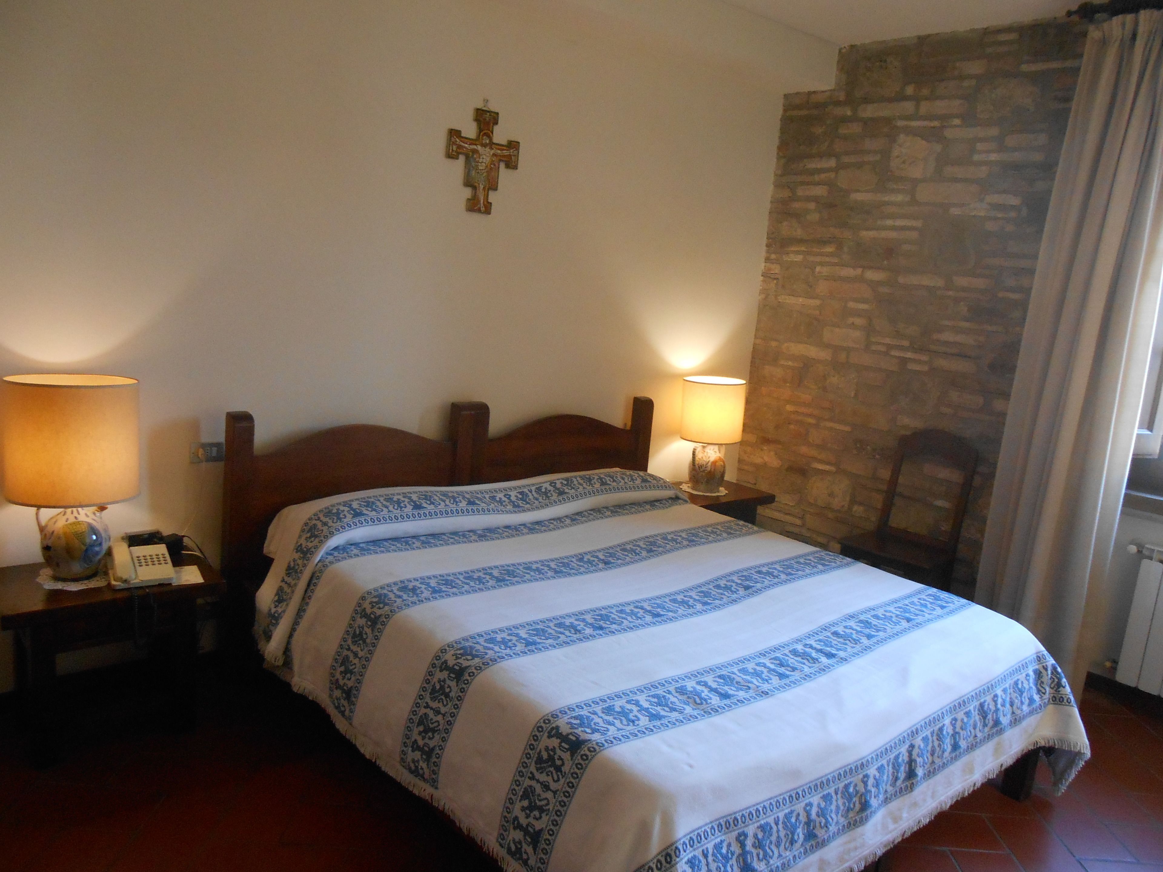 A bedroom with a wooden bed, two bedside lamps, a cross on the wall, and a stone wall.