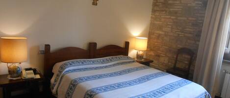 A bedroom with a wooden bed, two bedside lamps, a cross on the wall, and a stone wall.