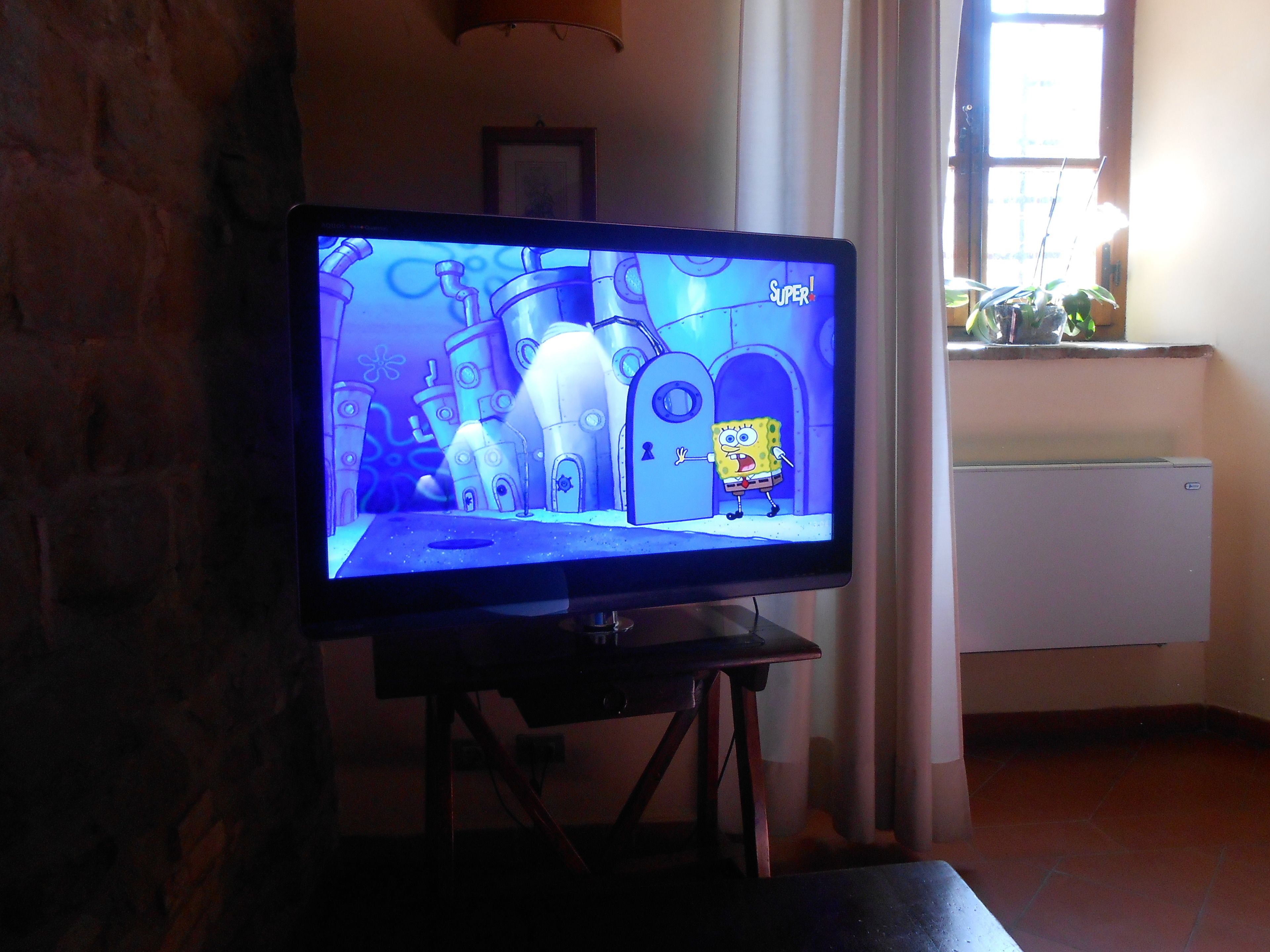 Un televisor muestra un programa animado, en una habitación con pared de ladrillo, una ventana con cortinas y un radiador.