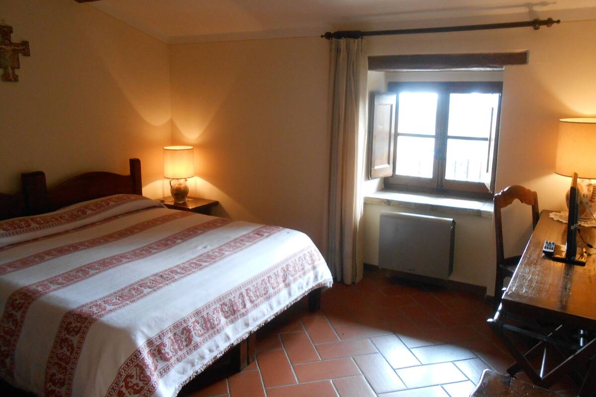 A bedroom with a bed, two bedside lamps, a window, a radiator, and a wooden table with a chair.
