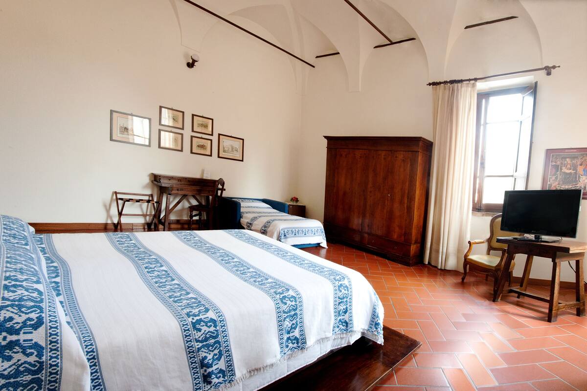 A hotel room with two beds, a wooden wardrobe, a desk with a television, and a window with curtains.