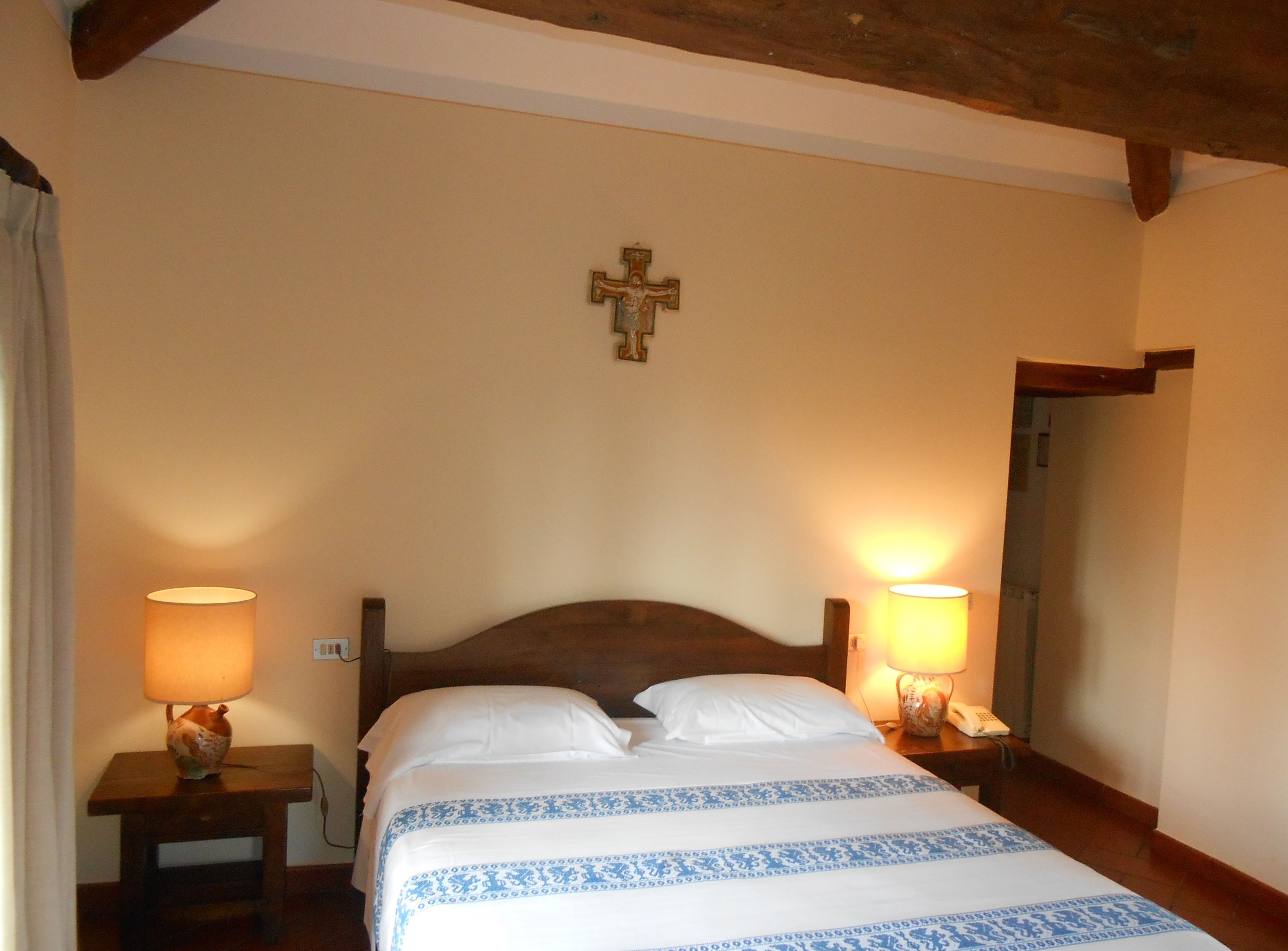 Hotelkamer met een houten hoofdbord, een kruis aan de muur, twee bedlampen en een bed met een blauw-wit gebederoppervlak.