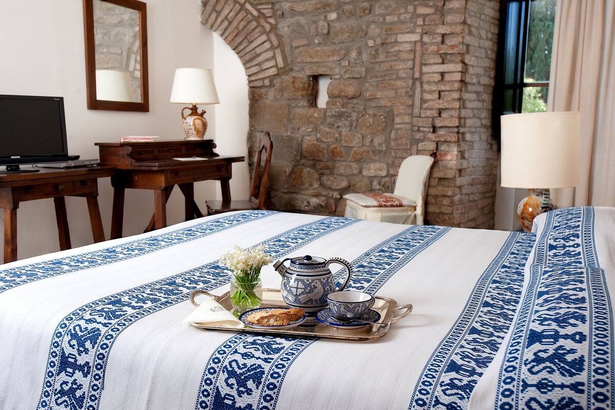 A neatly made bed with a blue and white patterned bedspread, a tray with a teapot, cup, and cookies, a laptop on a desk, and a stone wall.