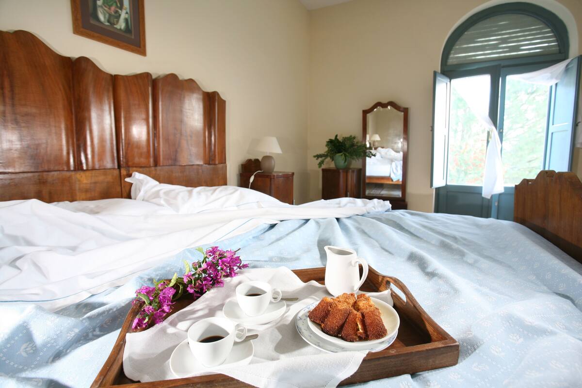 A neatly made bed with a tray of food and drinks, a wooden headboard, and a window with curtains.
