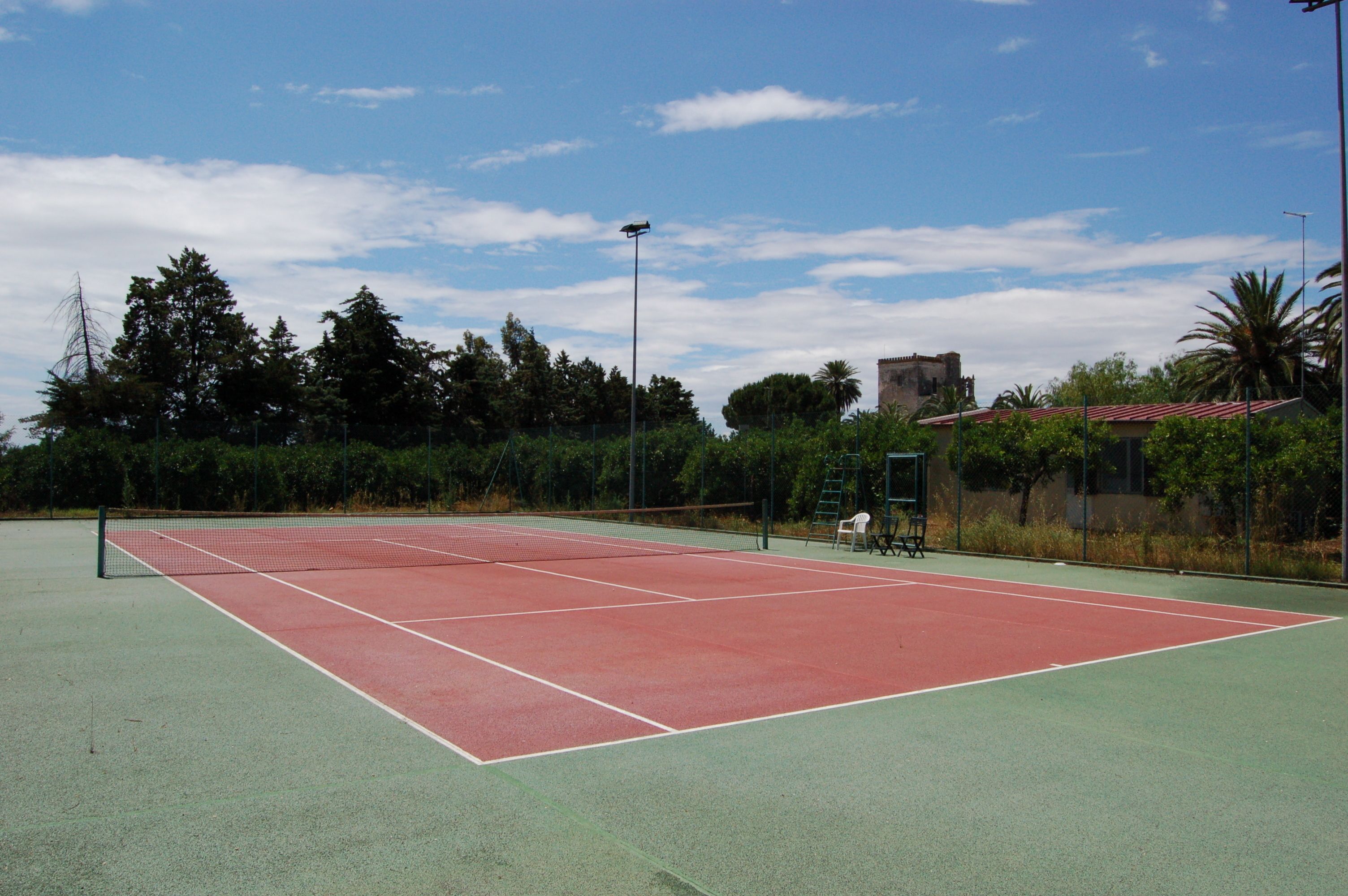 An outdoor tennis court with a net, surrounded by trees and a building with a red roof.