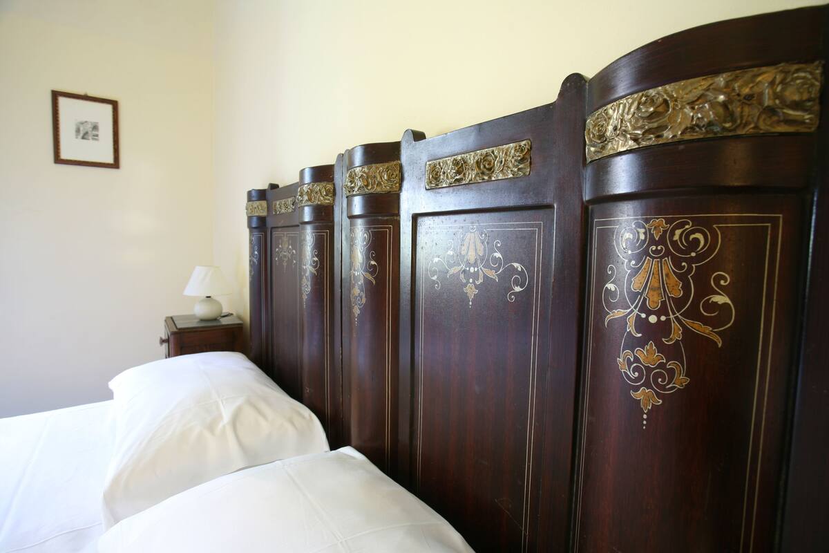 A neatly made bed with white linens in a room with a dark wooden headboard featuring intricate gold designs.