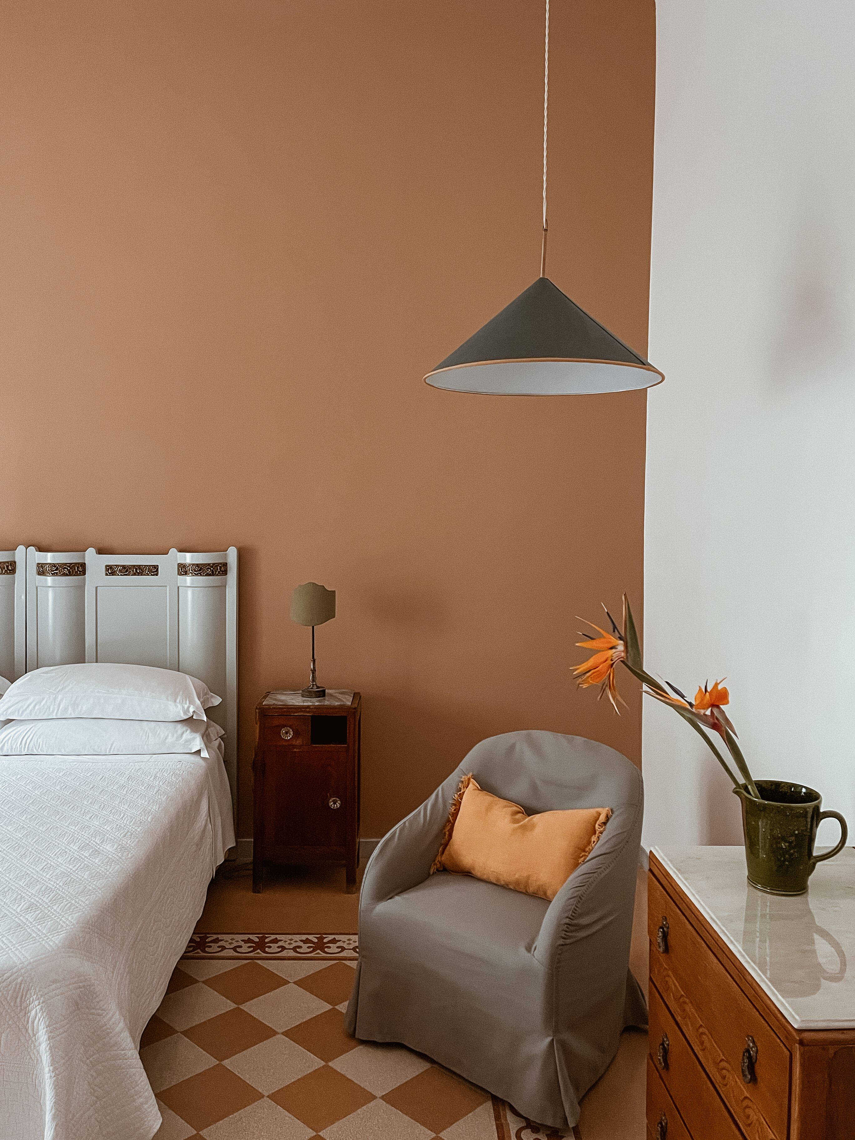 A bedroom with a bed, a chair, a nightstand, a lamp, and a vase with flowers.