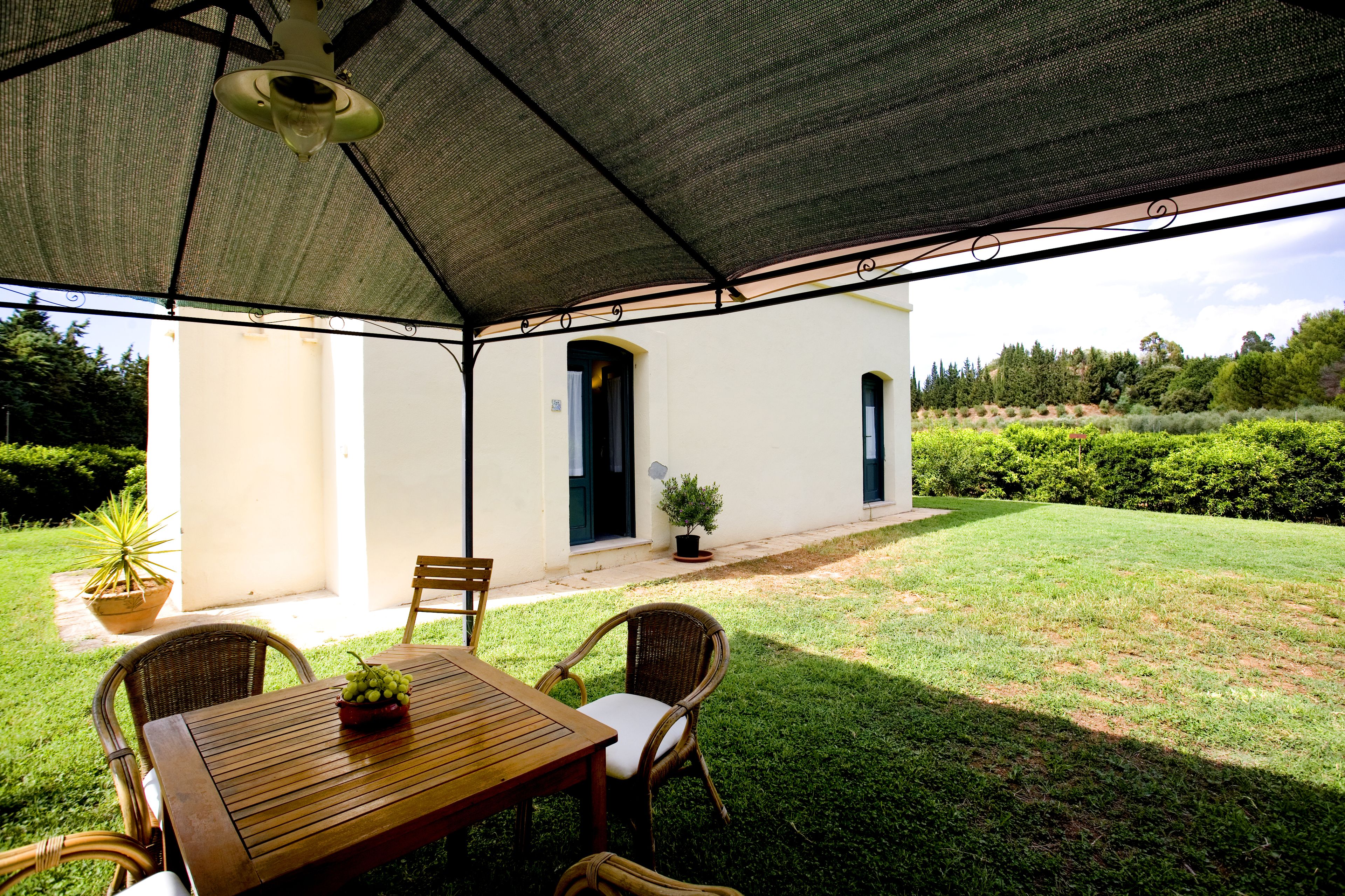 An outdoor patio with a wooden table and wicker chairs, a covered area with a ceiling fan, and a view of a white building and greenery.