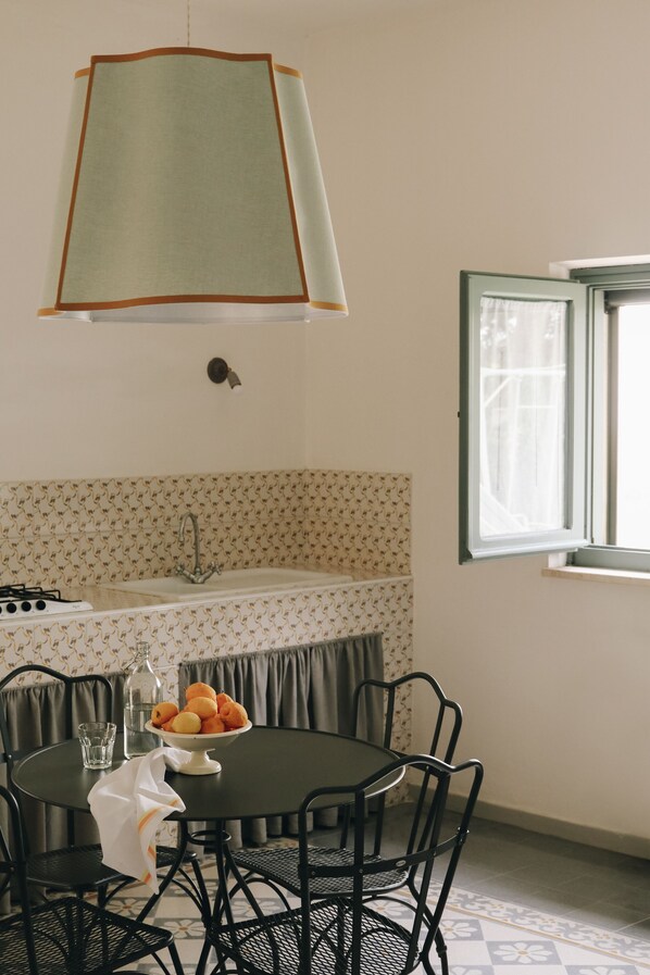 Un área de comedor con una mesa redonda, sillas, un cuenco de fruta, una lámpara colgante, una ventana y una zona de cocina con un lavabo y grifo.