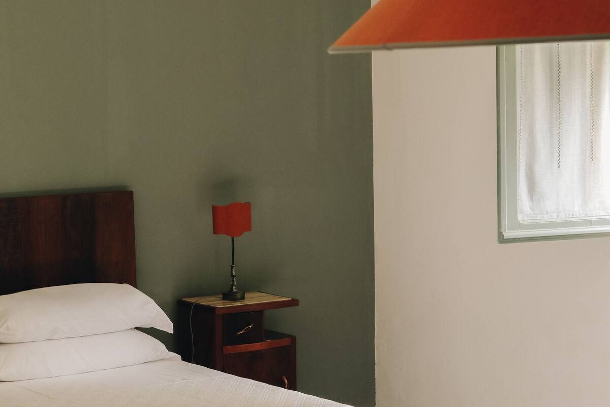 A bedroom with a bed, a nightstand, a red lamp, and a window with curtains.