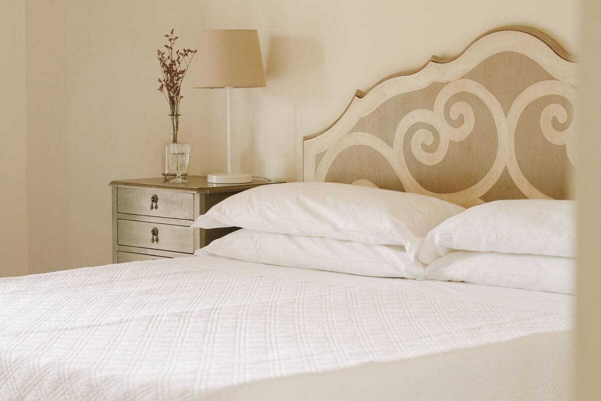 A neatly made bed with a headboard, a bedside table with a lamp, and a vase with flowers.