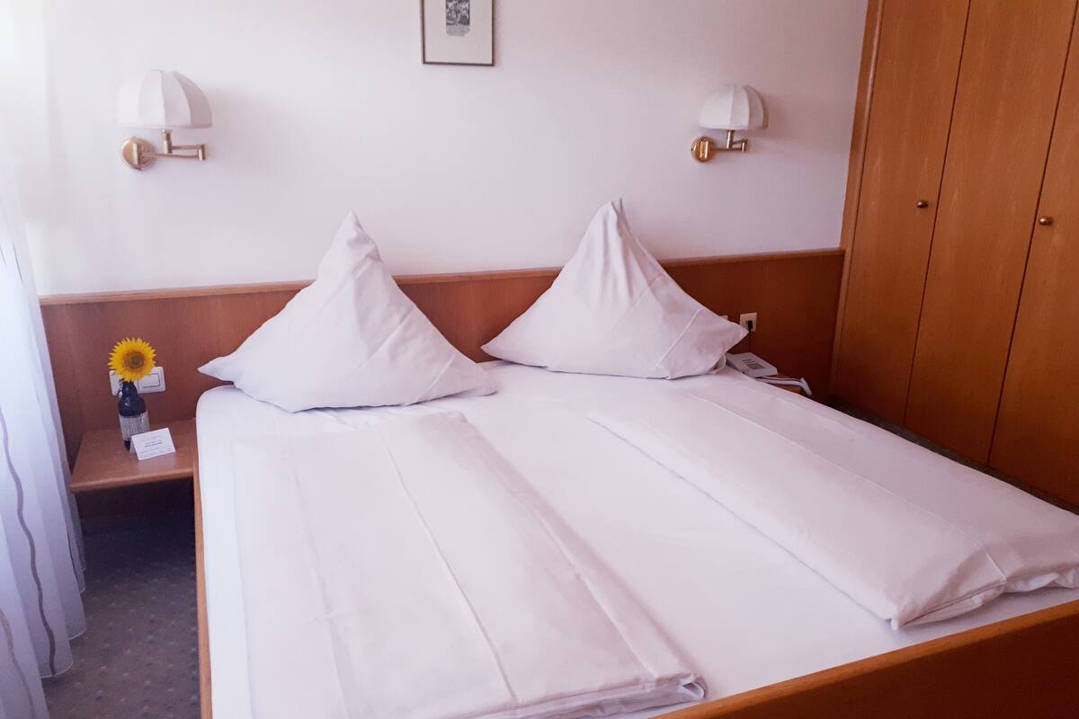 Een net opgemaakt bed met witte lakens, een houten hoofdbord en twee wandlampen.