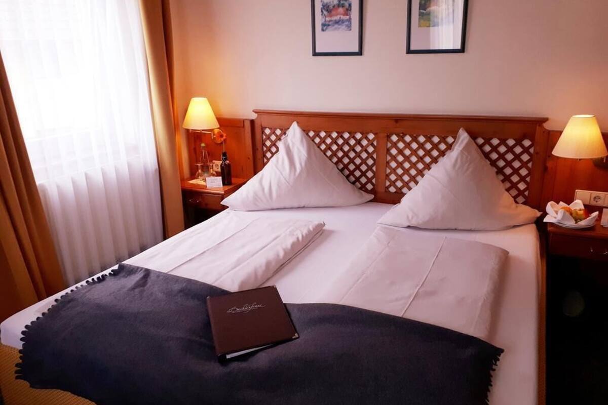Een net opgemaakt bed met witte lakens, twee kussens, een donkere plaid en een bruin boek. Er staan twee nachtlampjes, een houten hoofdbord met een spiegelmotief, en twee ingelijste foto’s aan de muur.