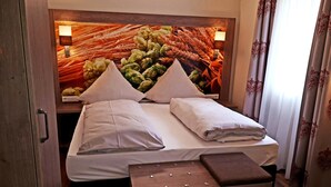 A hotel room with a bed featuring a headboard with a decorative wall art of artichokes and wheat. There is a bedside table with a lamp and a small bench at the foot of the bed. - Hotel Dachsenfranz (Zuzenhausen)