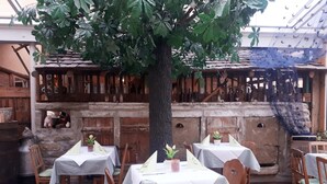 An indoor dining area with wooden tables and chairs, a large tree, and a stone wall with bottles. - Hotel Dachsenfranz (Zuzenhausen)