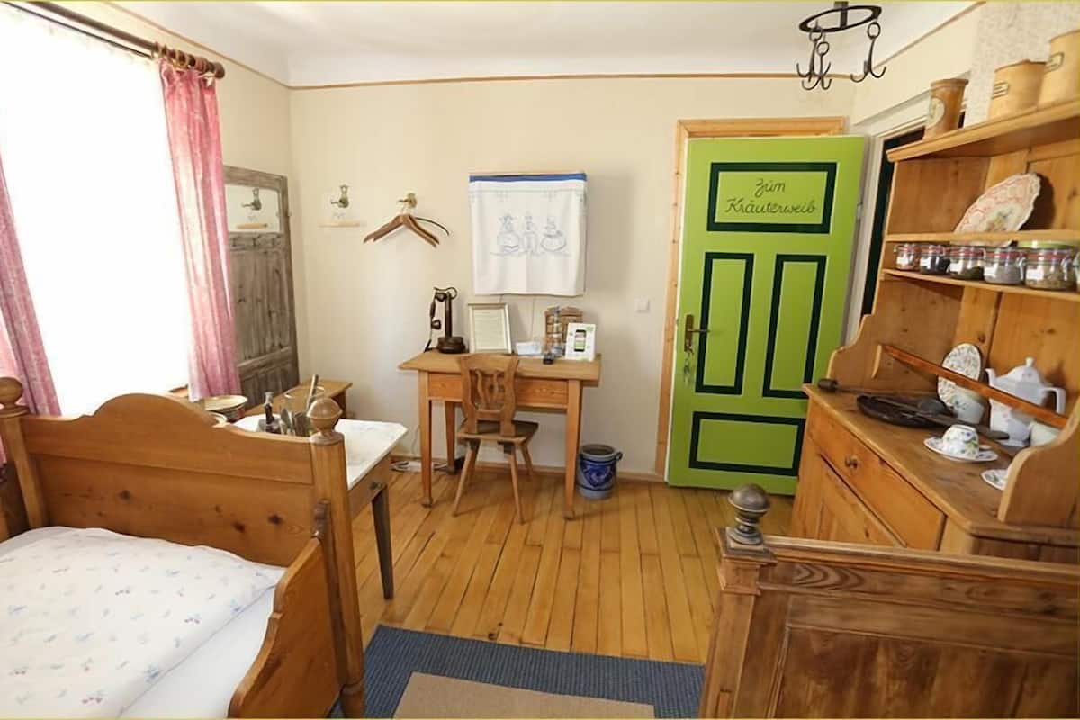 Een kamer met een houten bed, een bureau met stoel, een groene deur en planken met keukengerei.