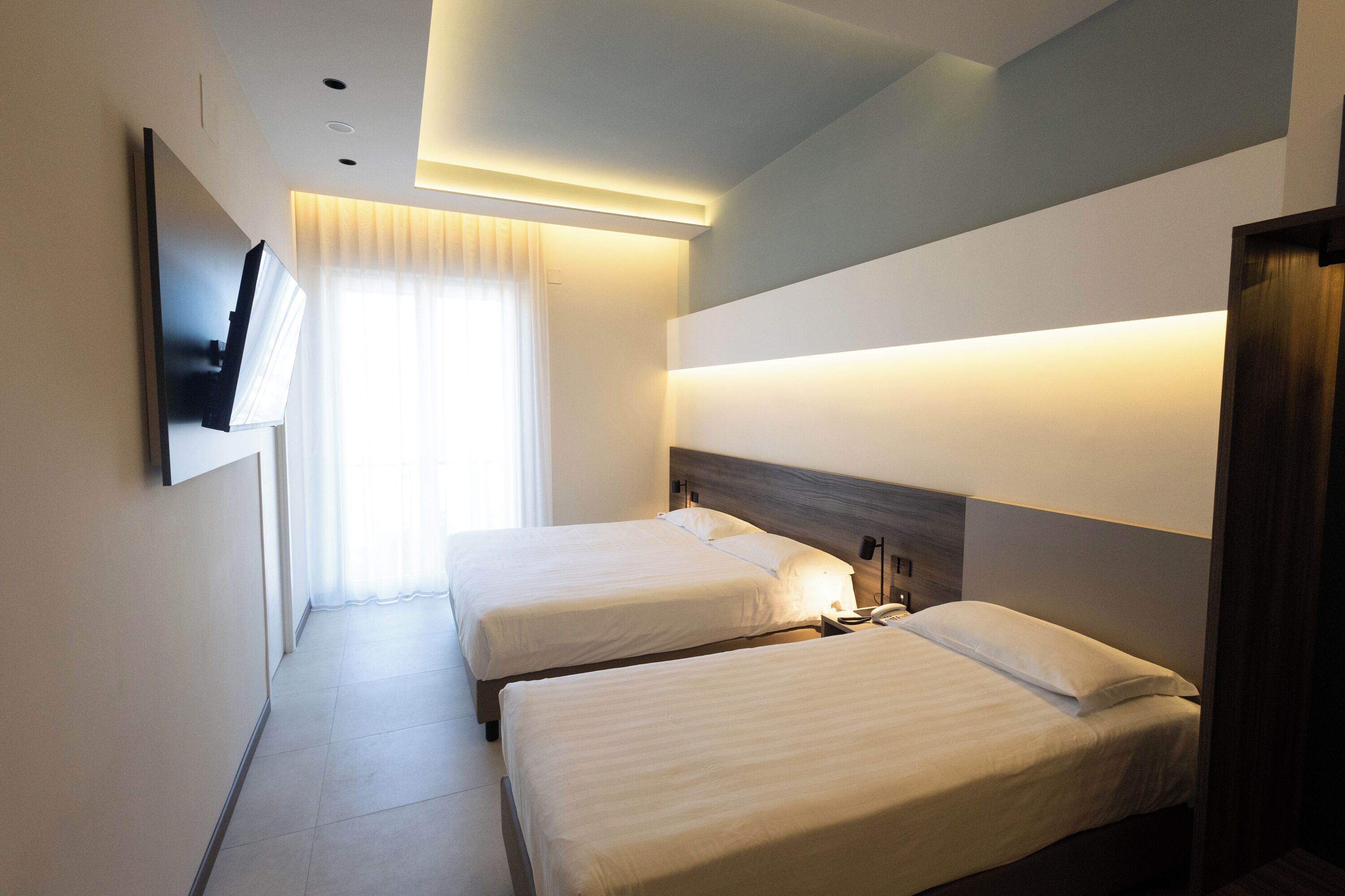 A hotel room with two beds, a flat-screen TV mounted on the wall, and a bedside table with a lamp.