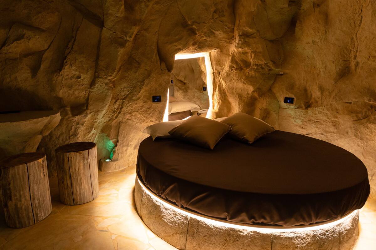 A circular bed in a cave-like room with a stone floor, wooden benches, and a small table.