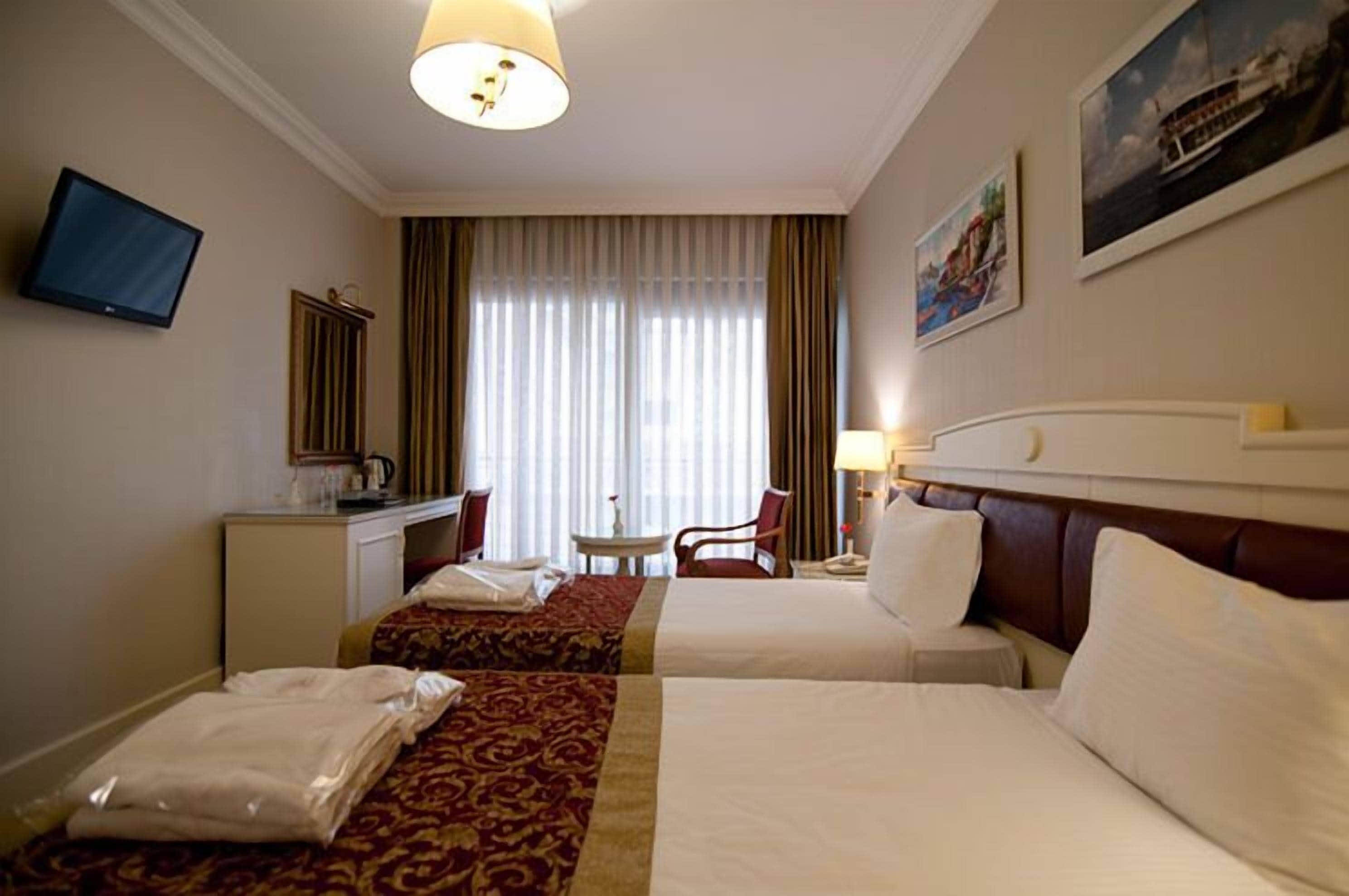 Quarto de hotel com duas camas, uma escrivaninha, uma cadeira, uma televisão e uma janela com cortinas.