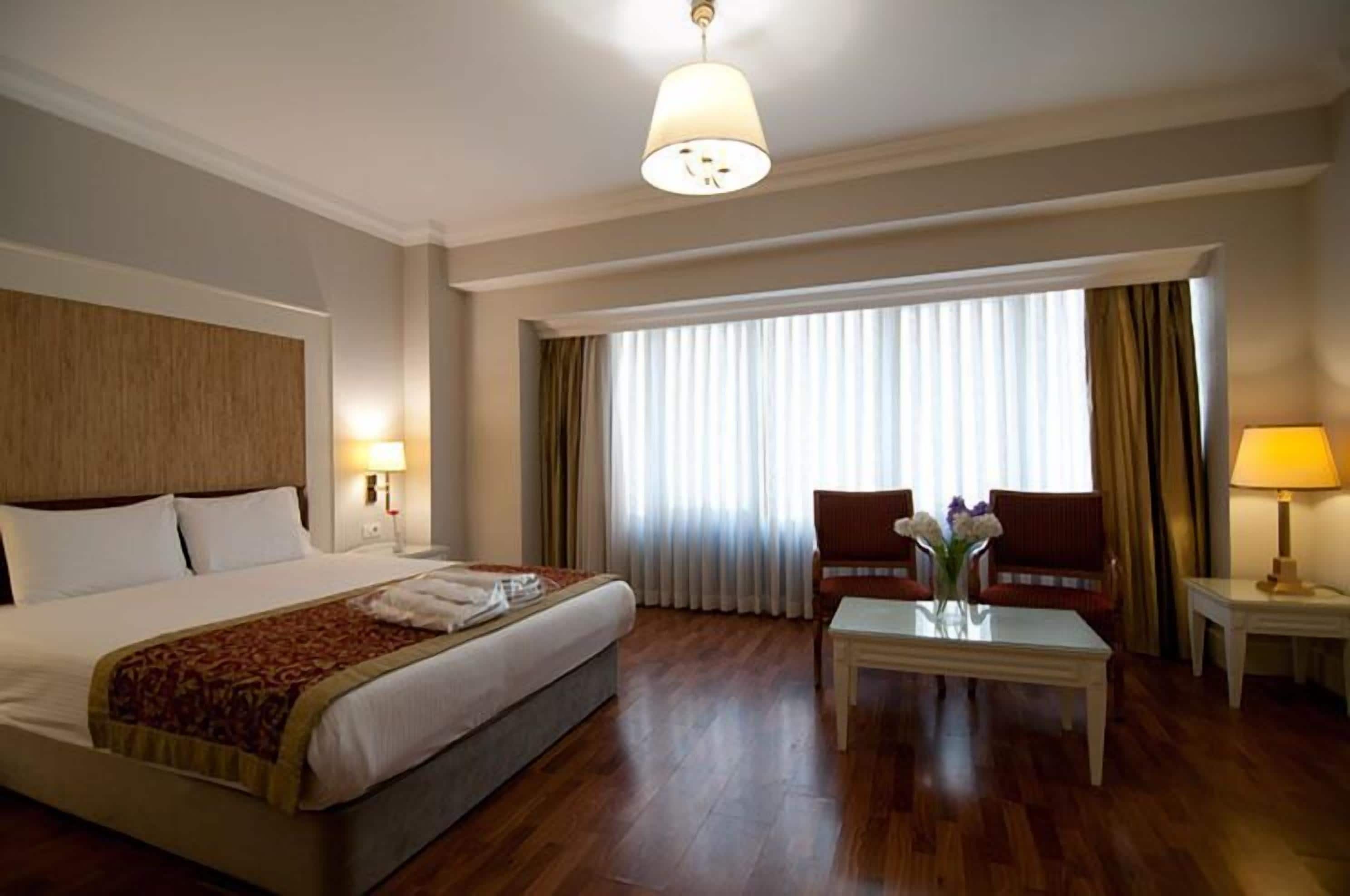 A hotel room with a bed, two armchairs, a coffee table, a lamp, and a large window with curtains.