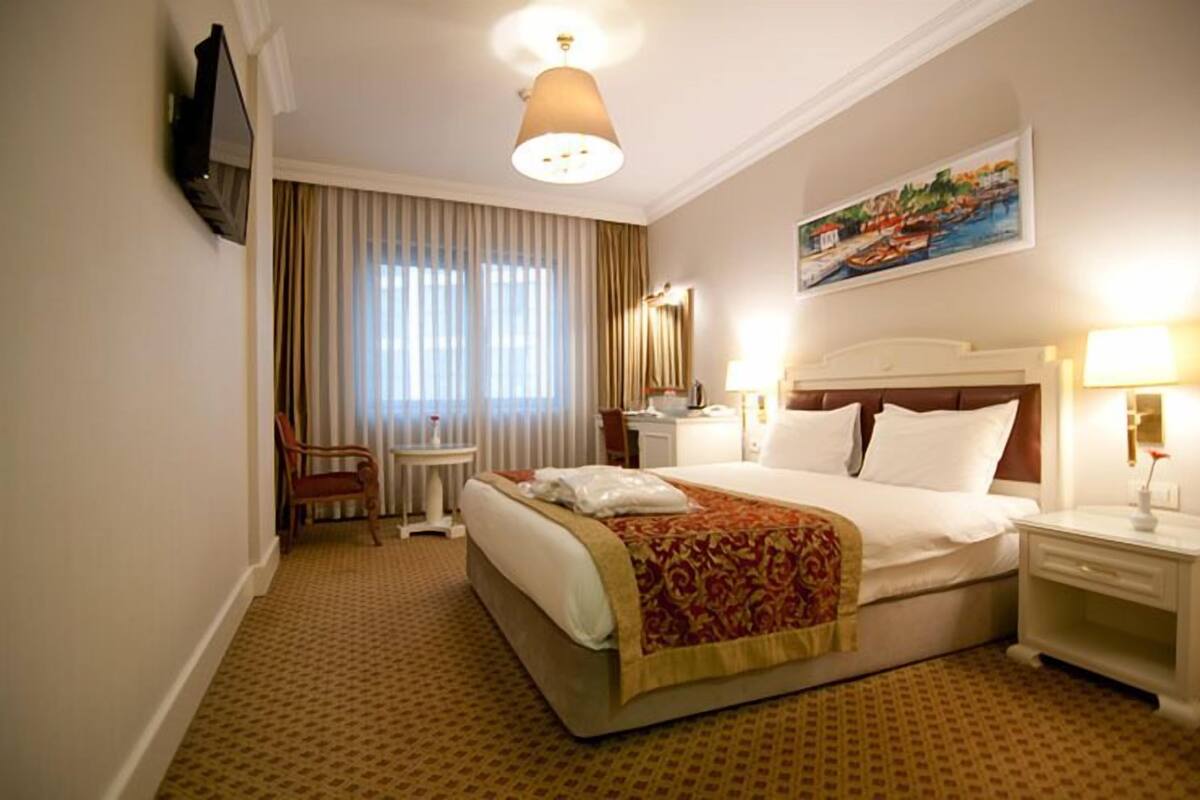 A hotel room with a bed, a chair, a desk, a television, and a painting on the wall.