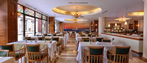 A spacious restaurant with multiple dining tables and chairs, a bar area, and large windows with a city view.
