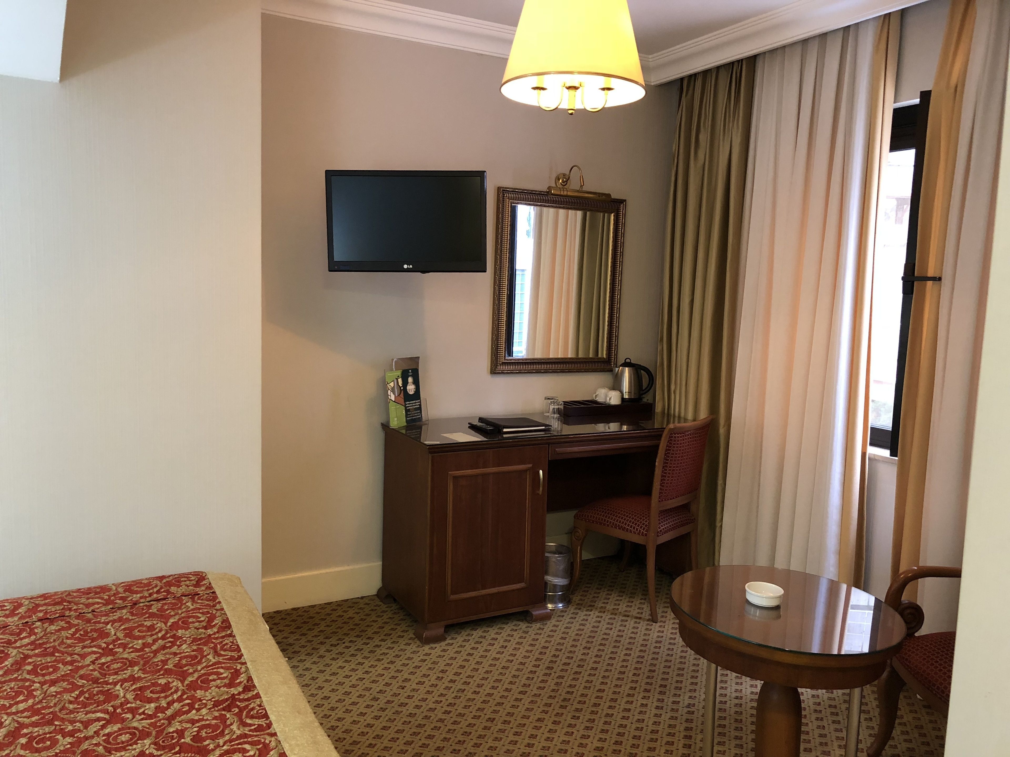 Une chambre d’hôtel comprenant un lit, un bureau, une chaise, une télévision, un miroir, une table et une fenêtre avec des rideaux.