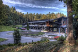 A modern building with a covered entrance, surrounded by a landscaped area with a circular driveway, trees, and a parking lot with several cars.