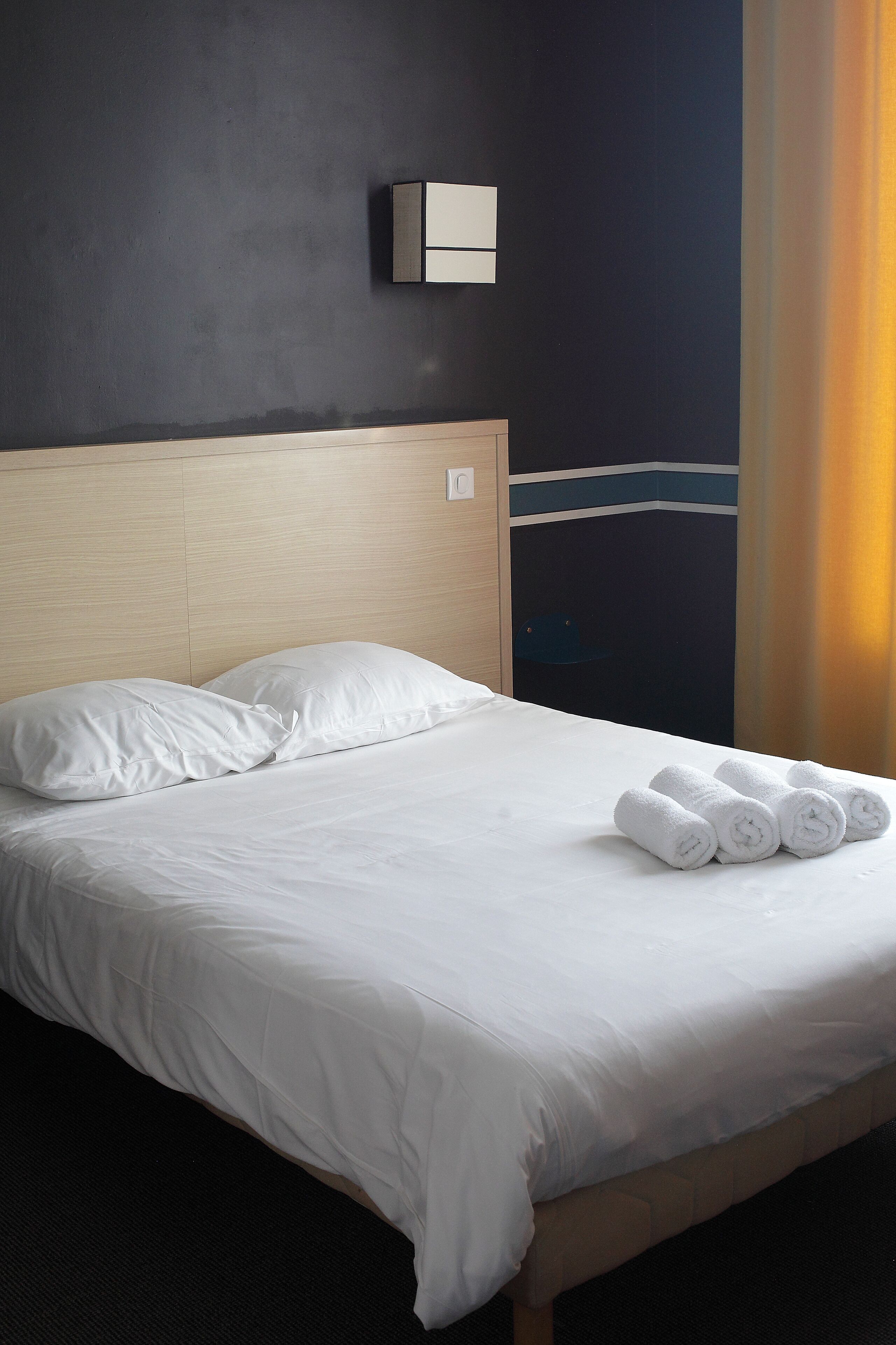 Cama bem arrumada com lençóis brancos e toalhas enroladas, cabeceira de madeira e luminária fixada na parede.