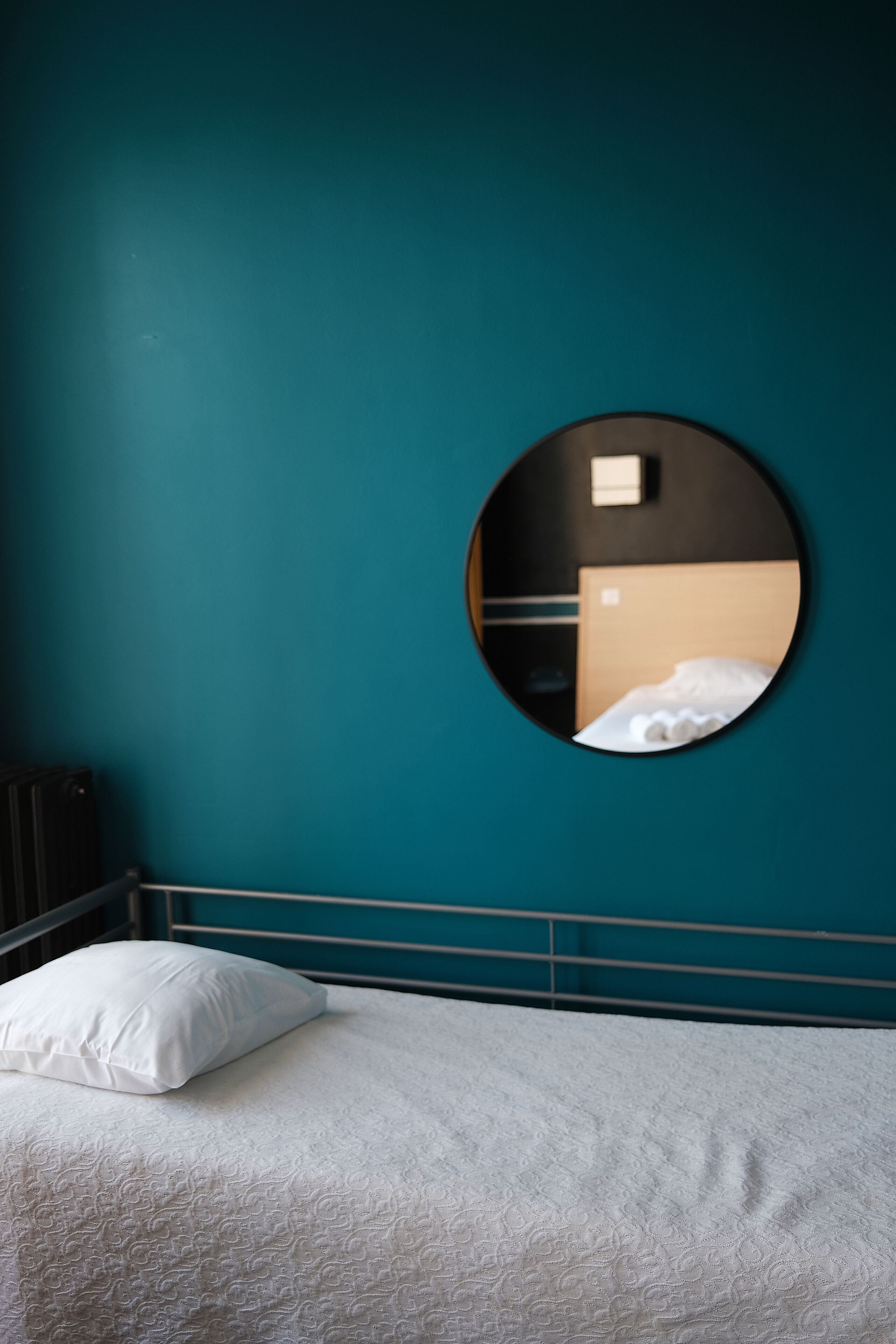 A bed with a white pillow and a textured blanket, a round mirror on the wall, and a teal-colored wall.