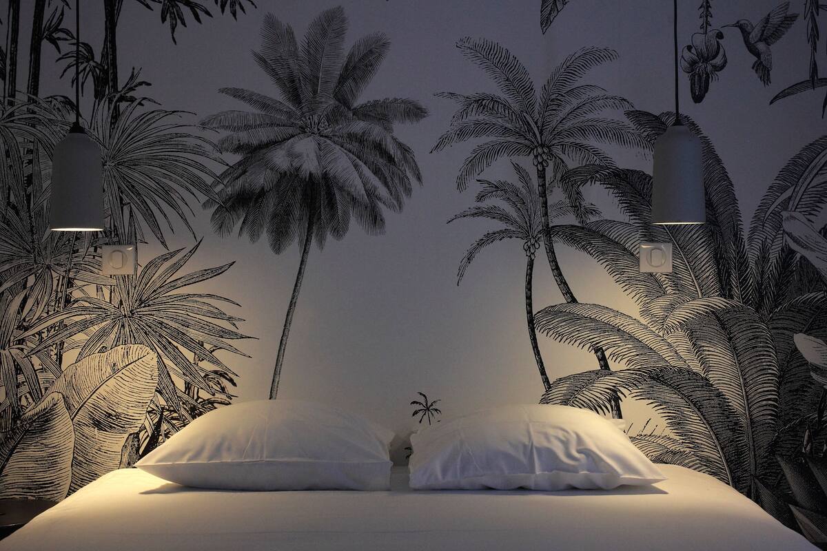 A bedroom with a bed, white pillows, and a wall mural of palm trees.