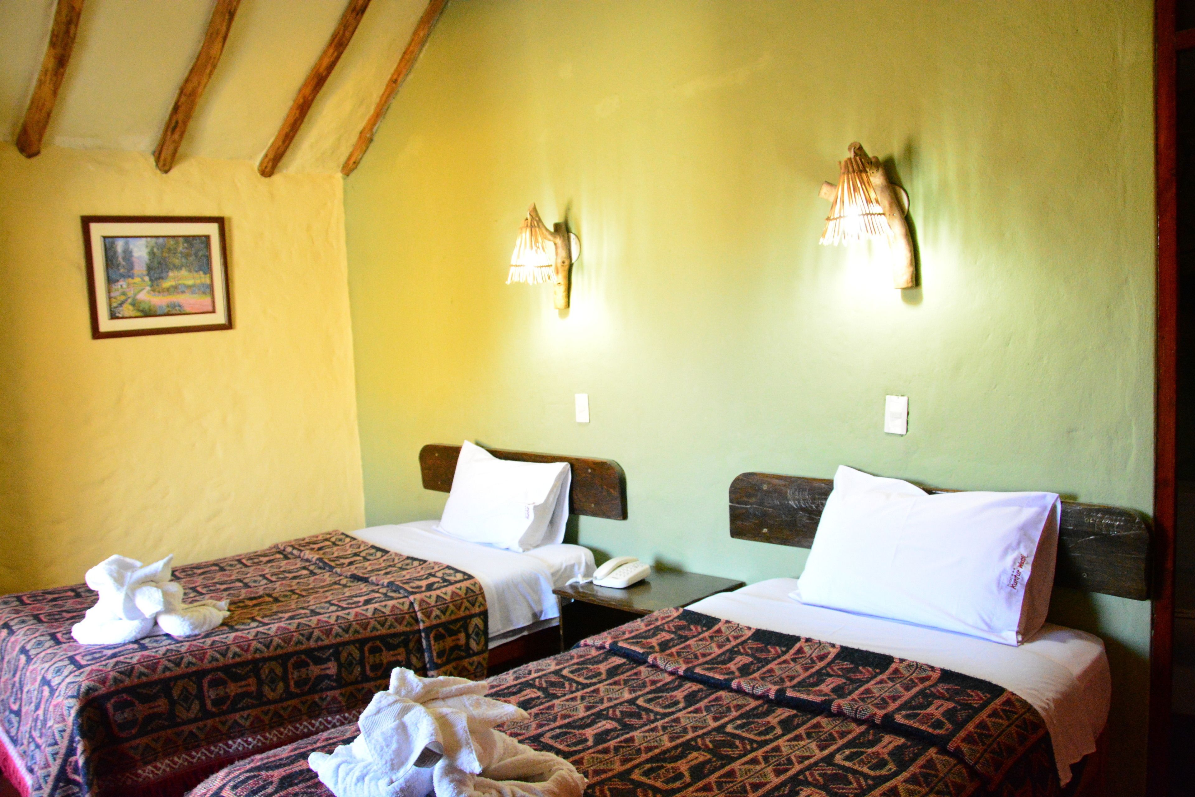 A room with two beds, each with patterned bedding and white pillows. There are wall-mounted lights and a painting on the wall.