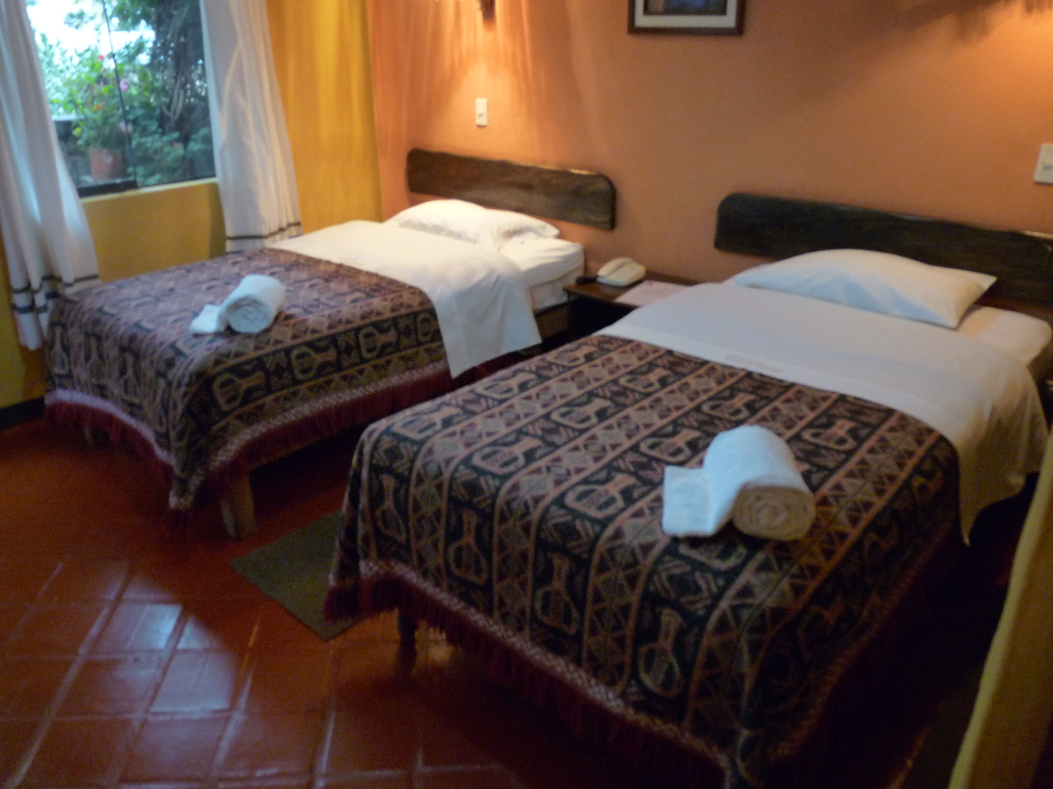 Two double beds with patterned bedspreads and matching pillows, a bedside table with a phone, and a window with curtains.