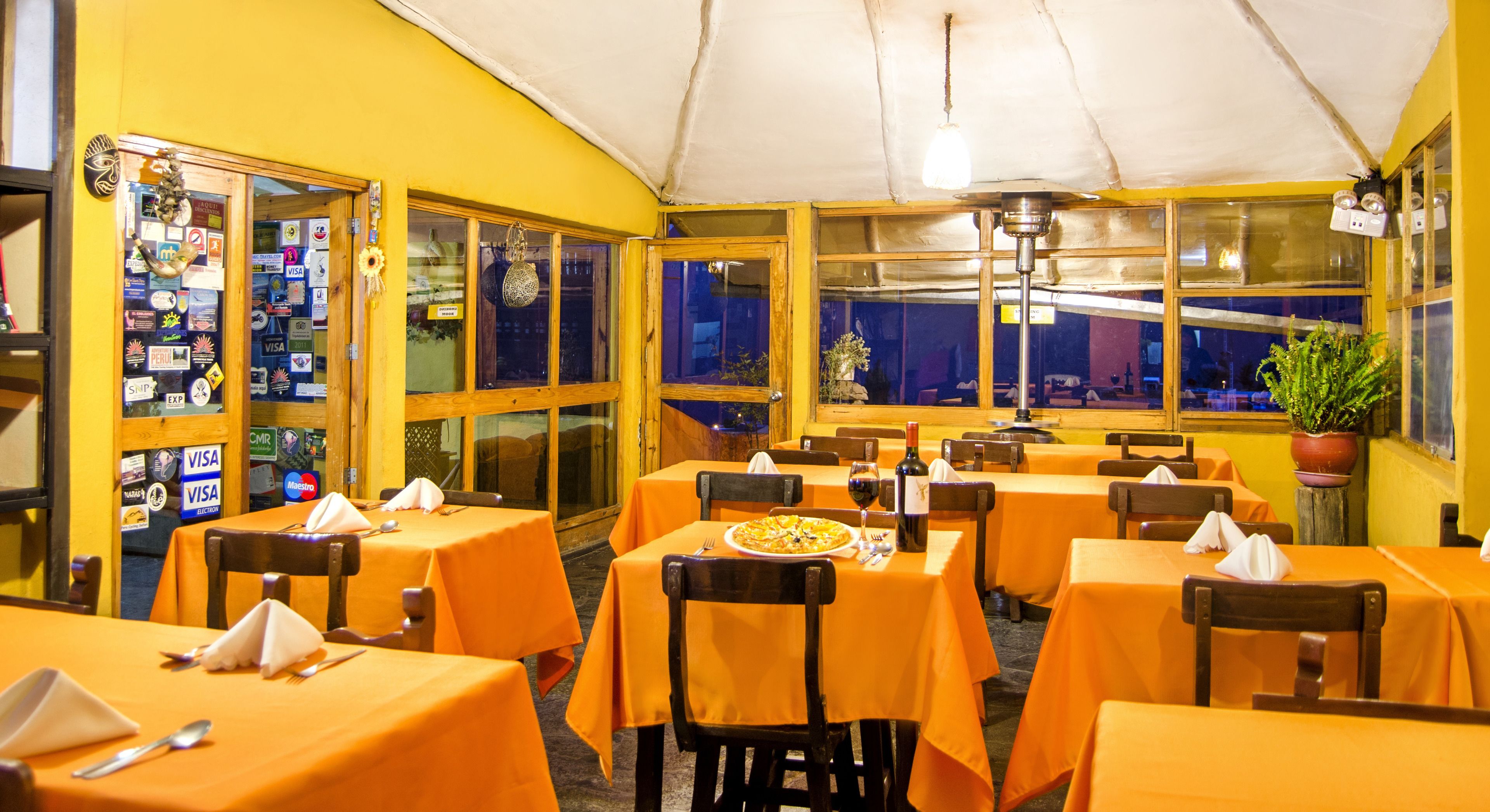 Un restaurant où les tables sont recouvertes de nappes orange, où les chaises sont en bois, et où une grande fenêtre offre une vue sur le ciel nocturne.