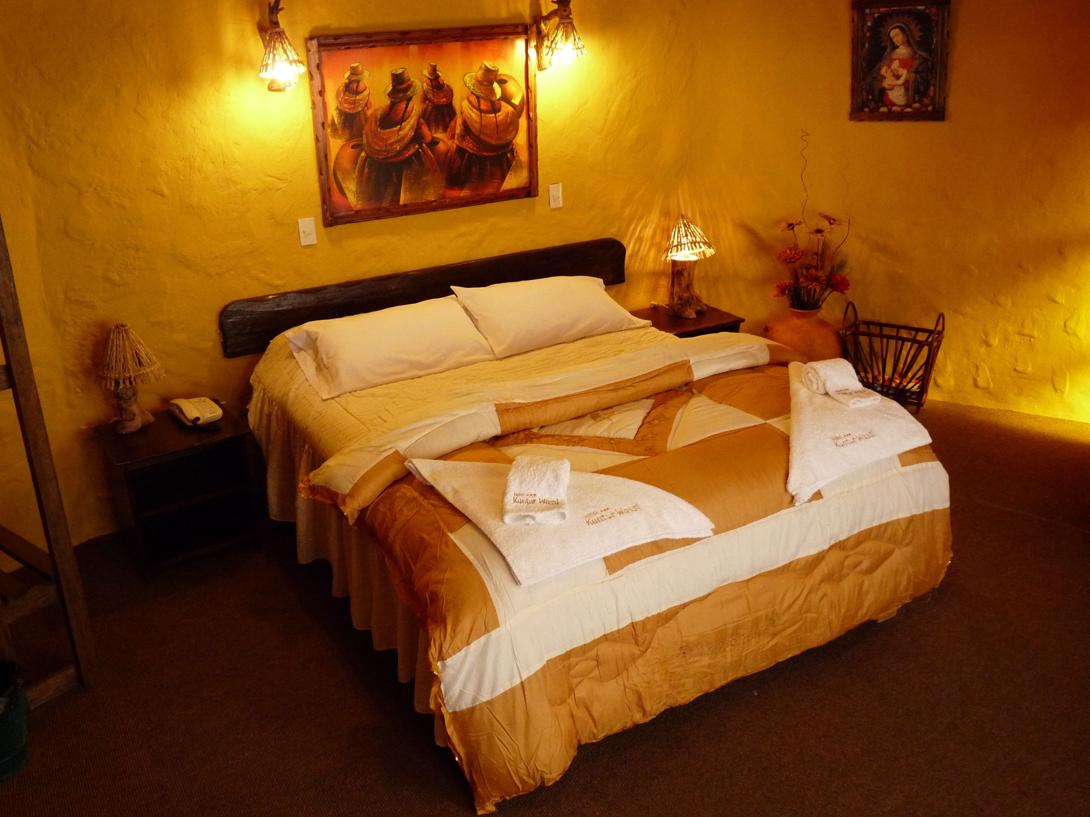 A hotel room with a large bed, a nightstand, a lamp, a painting on the wall, and a small table with a vase of flowers.