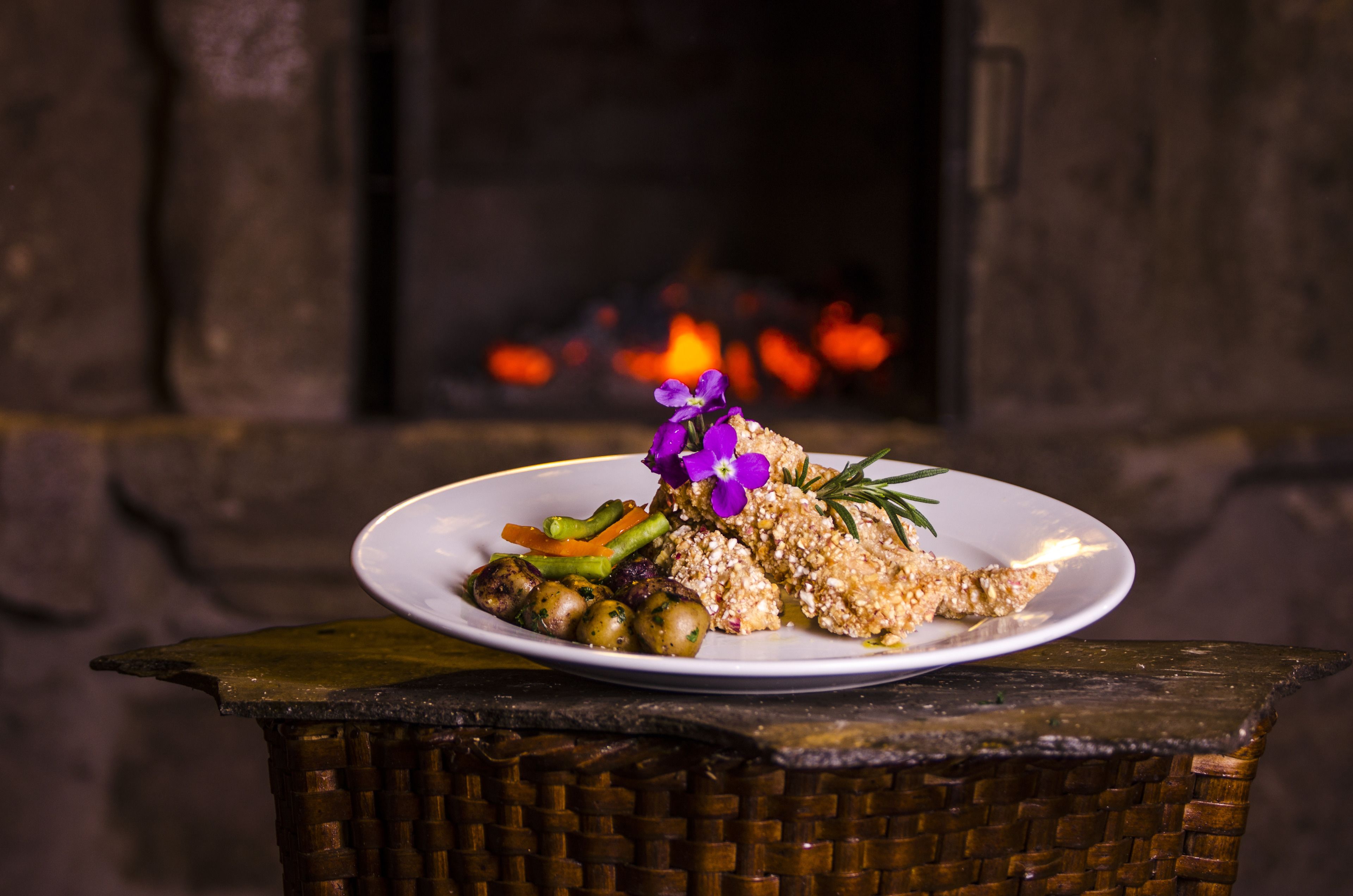 Un plato de comida con verduras y una flor morada sobre una mesa frente a una chimenea.