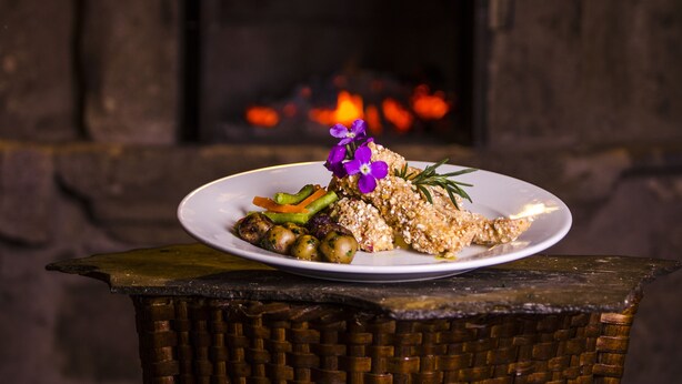 Un plato de comida con verduras y una flor morada sobre una mesa frente a una chimenea.