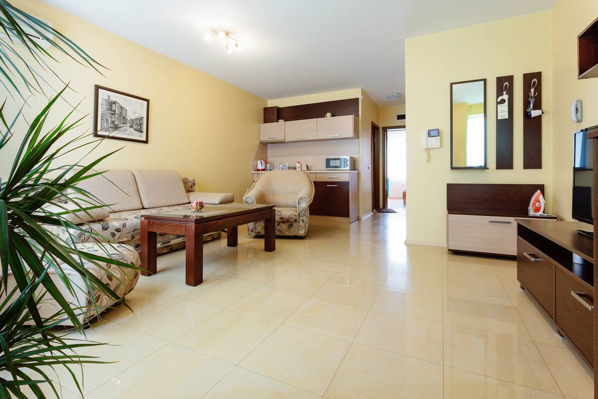A living room with a sofa, a coffee table, and a chair. There is a kitchen area with cabinets and a microwave.