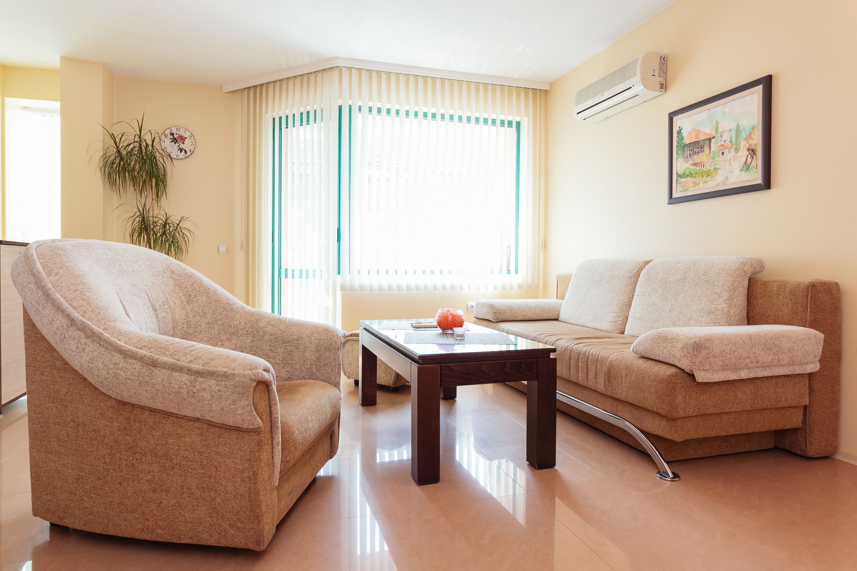 A living room with a beige sofa, a beige armchair, a glass-top coffee table, and a potted plant.