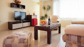 A living room with a TV, sofa, ottoman, and a coffee table with fruit and a bottle.