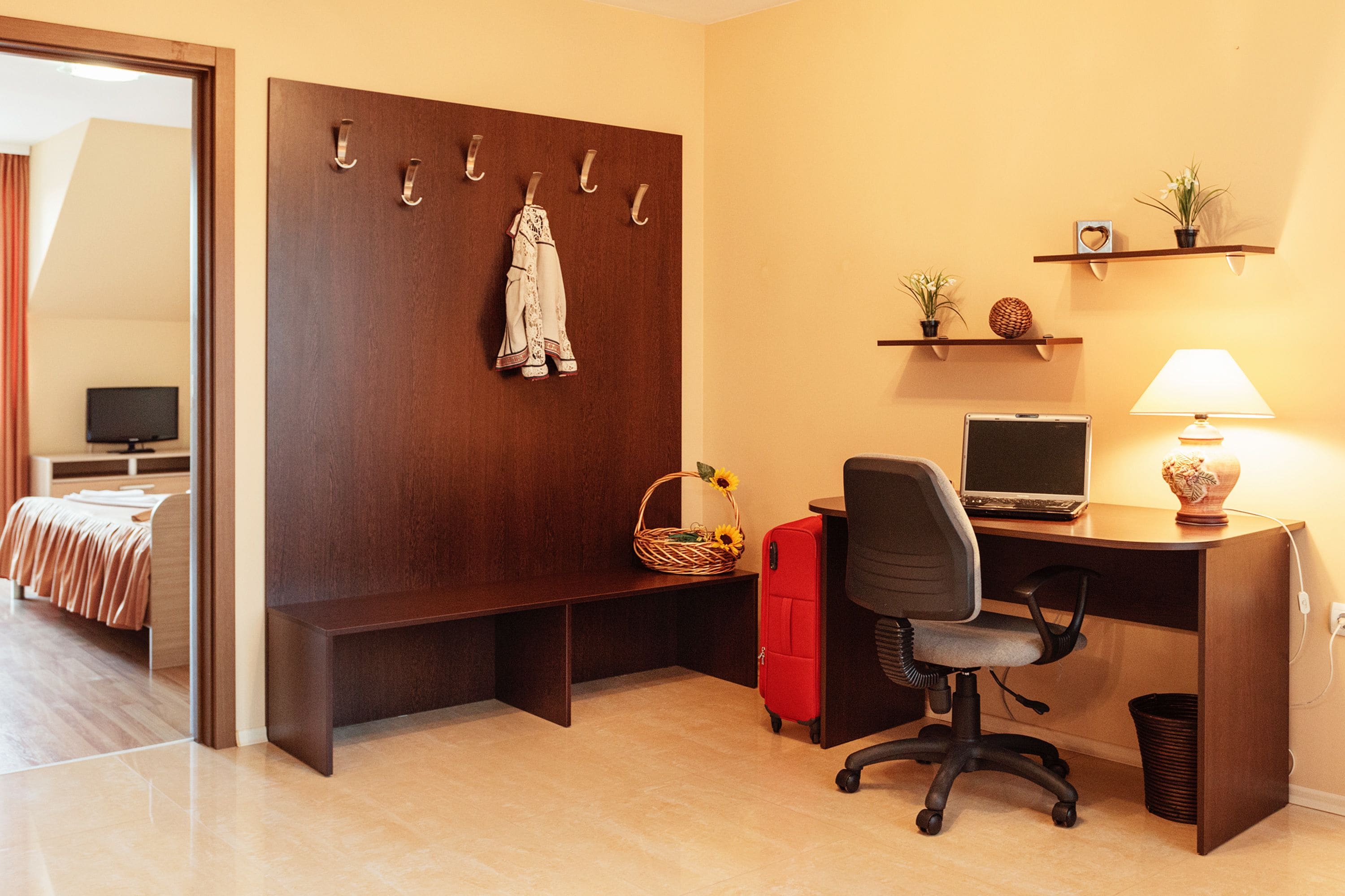 Una habitación de hotel con un escritorio de madera, una computadora, una lámpara, una maleta roja, una cesta de mimbre con girasoles, un perchero con un vestido y una cama con un televisor al fondo.