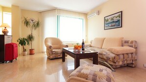 A living room with a sofa, armchair, and coffee table. There is a potted plant, a red suitcase, and a painting on the wall. - Bright House (Plovdiv)