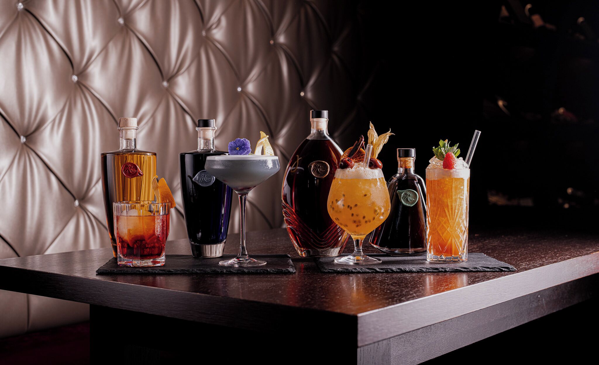 A bar setting with various bottles and glasses of drinks, a tufted leather wall, and a dark wooden counter.