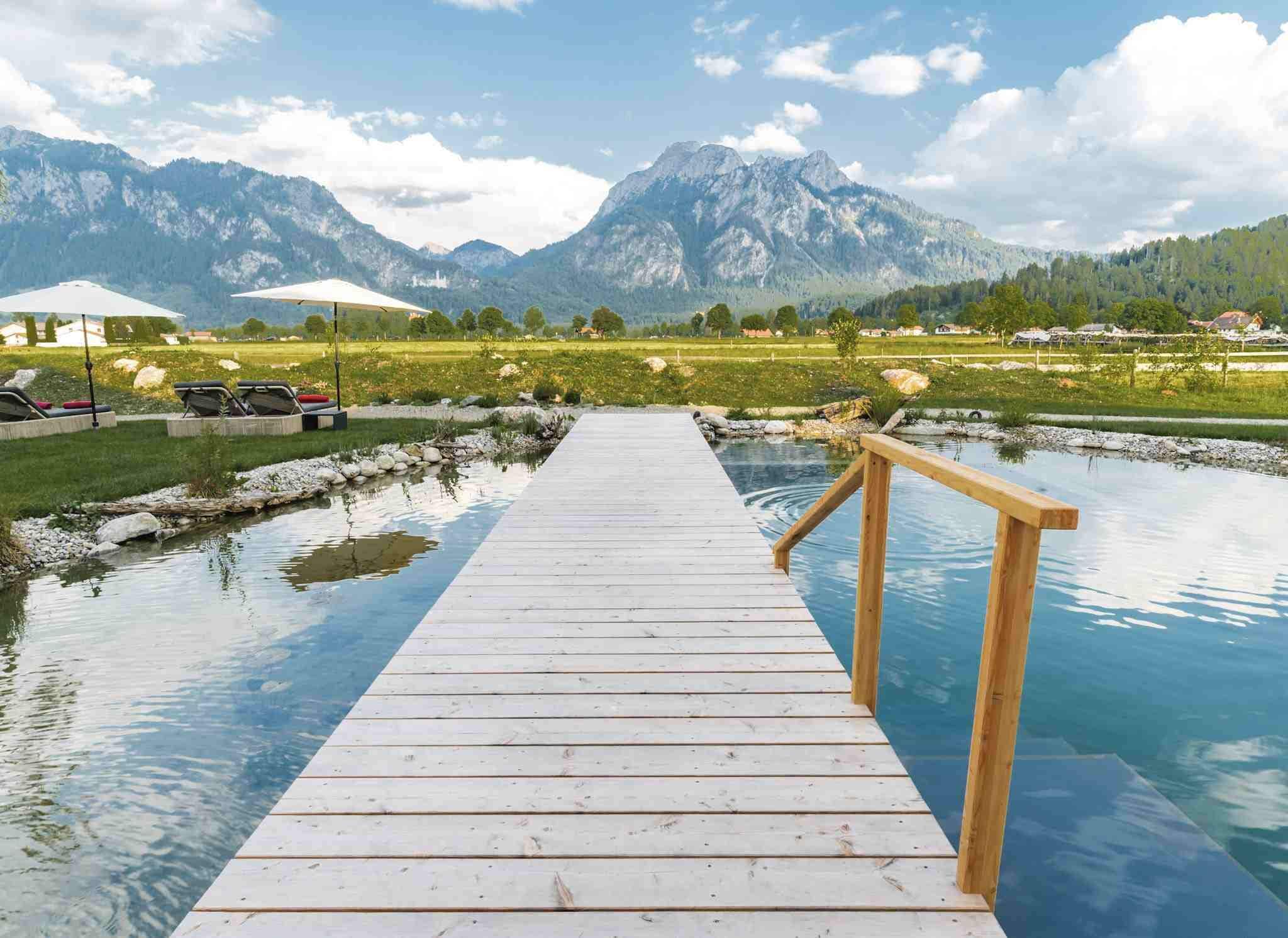 Un muelle de madera que conduce a una piscina cristalina con una montaña al fondo.
