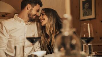 Un couple est assis à une table, devant des verres à vin et une bouteille, dans une pièce aux murs en bois et ornée d’un tableau encadré.