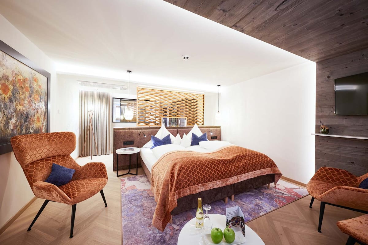 Une chambre d’hôtel moderne avec un grand lit, deux fauteuils, une petite table avec une bouteille et des pommes, et un téléviseur fixé au mur.